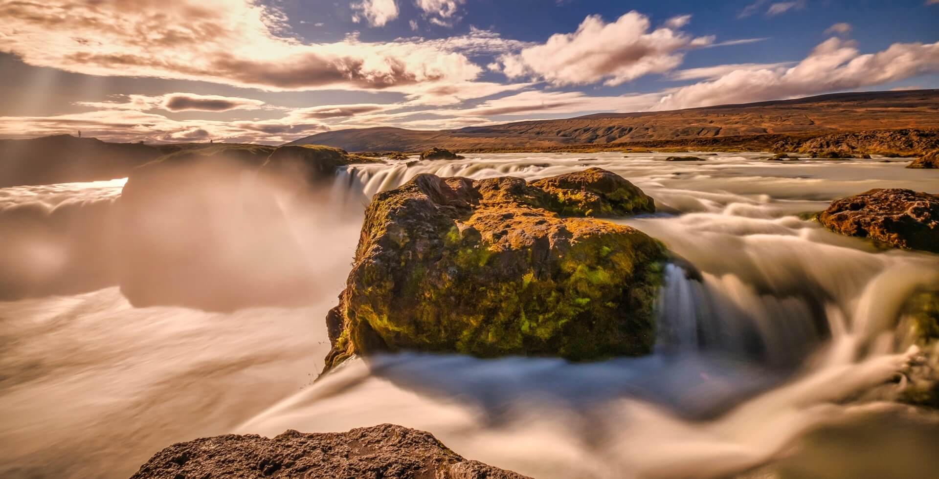 Footer Goðafoss in long exposer, blue sky, warm autumn colors in the Sun rays, a big rock in the middle